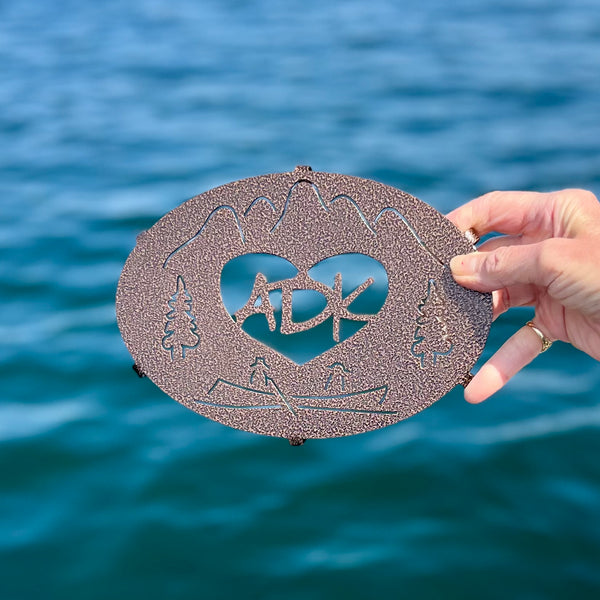 ADK in a heart with a mountain, trees and canoe scene on a copper penny metal trivet.