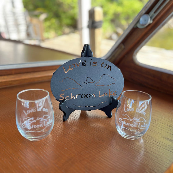 Black metal Love is on Schroon Lake trivet paired with two clear stemless wine glasses.
