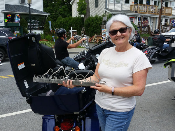 Love is in the Adirondacks text on the base of stainless sign inside the outline of the Adirondack mountain scape.  A customer is standing next to her motorcycle holding up her sign.