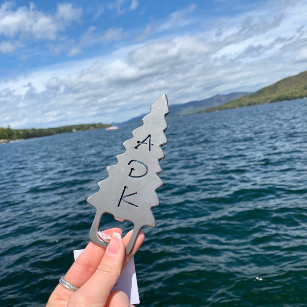 Handcrafted stainless steel bottle opener with a ADK cut out of an Adirondack evergreen tree. The bottle opener tab is at the base of the tree.