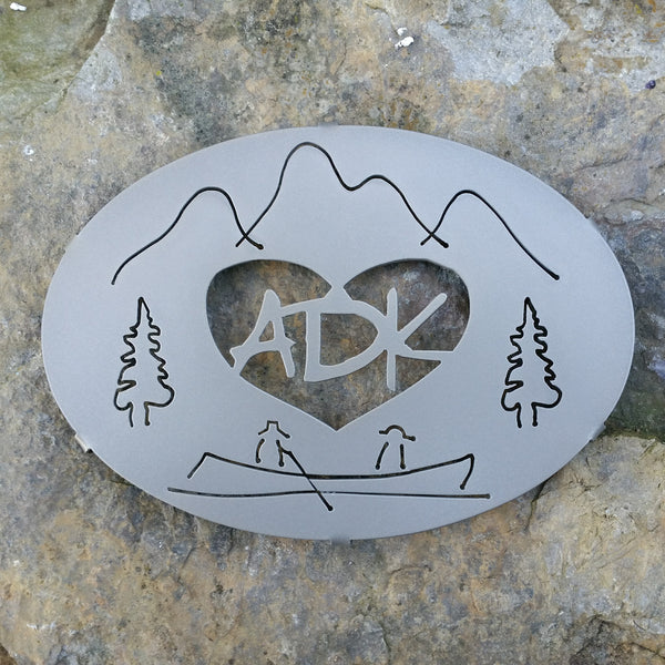 ADK in a heart with the Adirondack mountains, trees and guide boat. It is powder coated gold haze. It is 8" x 6".
