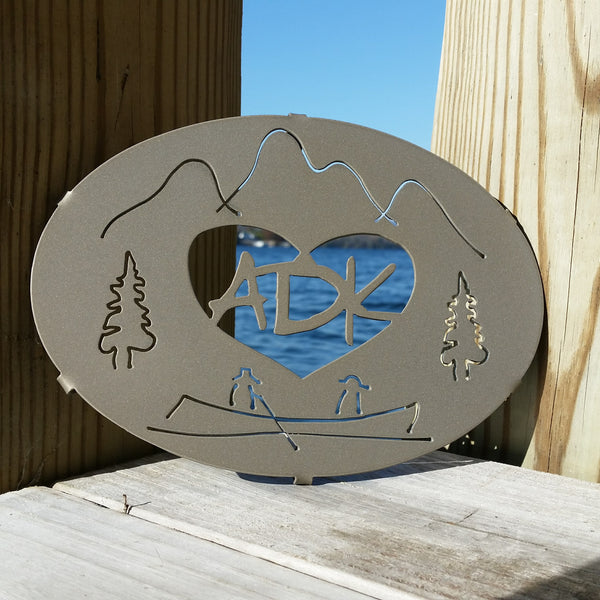ADK in a heart with the Adirondack mountains, trees and guide boat. It is powder coated gold haze. It is 8" x 6".