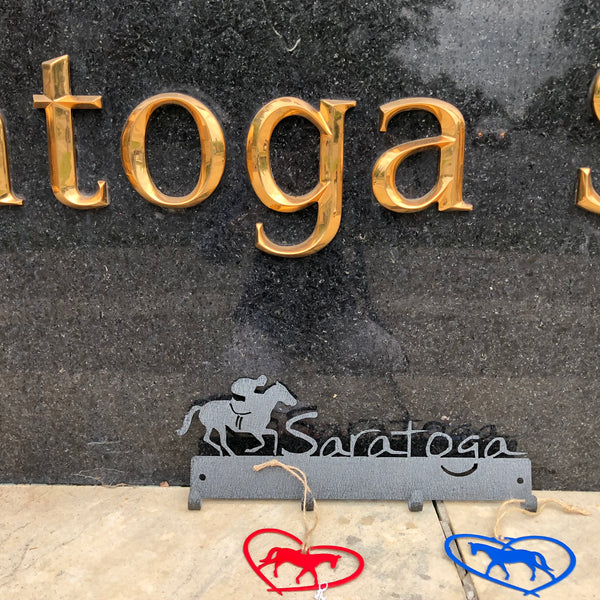 Jockey riding a horse with the word Saratoga. It is a metal 4 hook wall mount color black/graphite. Two horse ornaments (red and blue) are hanging off of the hooks.
