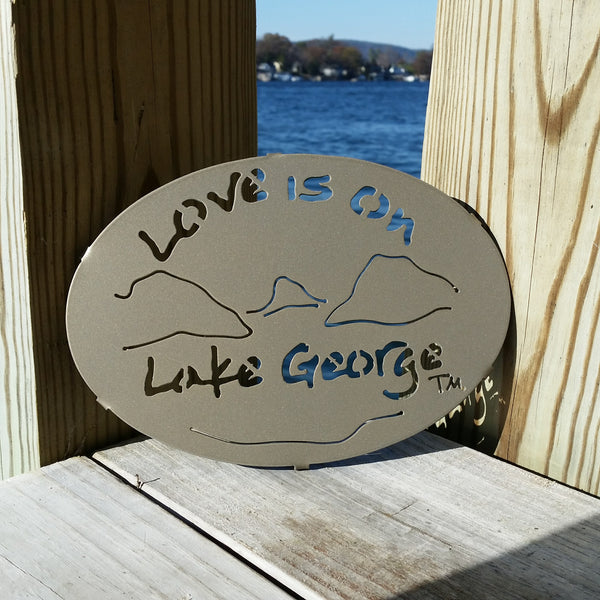 Text Love is on Lake George with the outline of the lake and surrounding Adirondack mountains on a metal trivet; color is gold haze