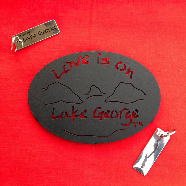 Love is on Lake George text over a lake/mountain scene on a black metal trivet. A stainless keychain with the silhouette of Lake George on one side and the text Love is on Lake George on the other side.