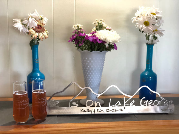 Love is on Lake George Text on a metal sign with the cutout of the Mountains.  Two Love is on Lake George stemless champagne flute.