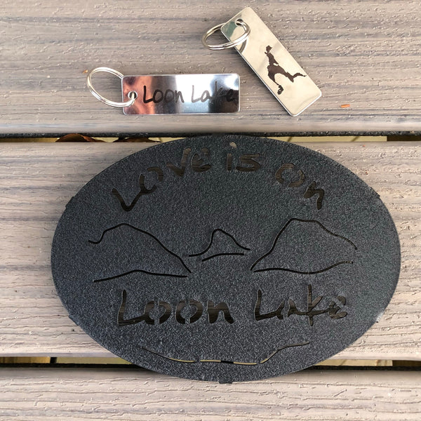 Love is on Schroon Lake text over/under a lake/mountain scene on a black metal trivet. It is paired with a stainless Schroon Lake keychain that has the silhouette of the lake on one side.