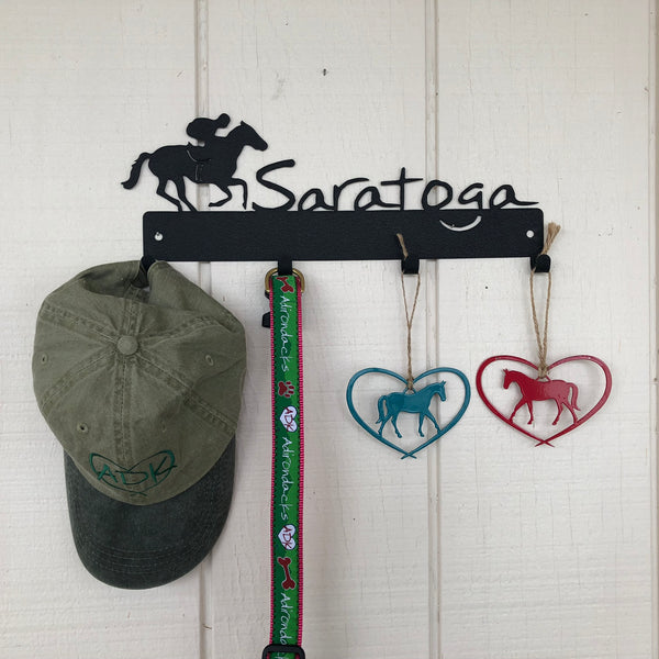 Jockey riding a horse with the word Saratoga. It is a metal 4 hook wall mount color black/graphite. A ball cap, dog collar and two horse ornaments are hanging off of the hooks.