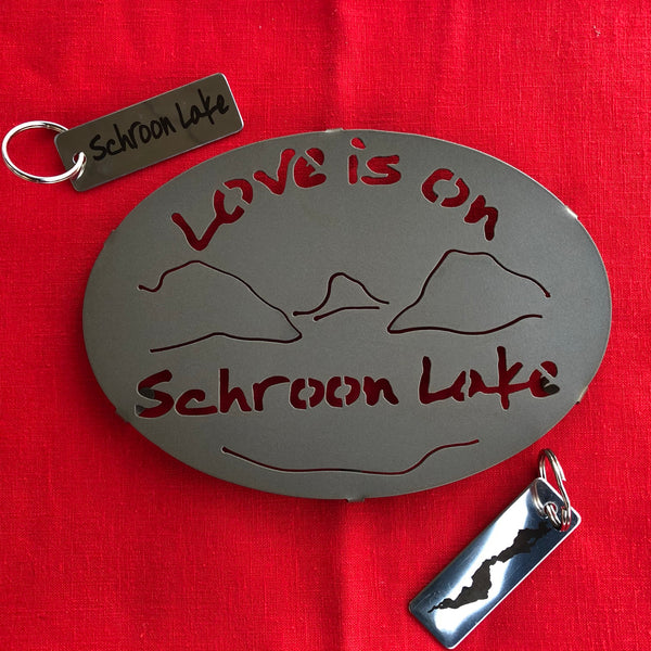 Love is on Schroon Lake text over/under a lake/mountain scene on a black metal trivet. It is paired with a stainless Schroon Lake keychain that has the silhouette of the lake on one side.