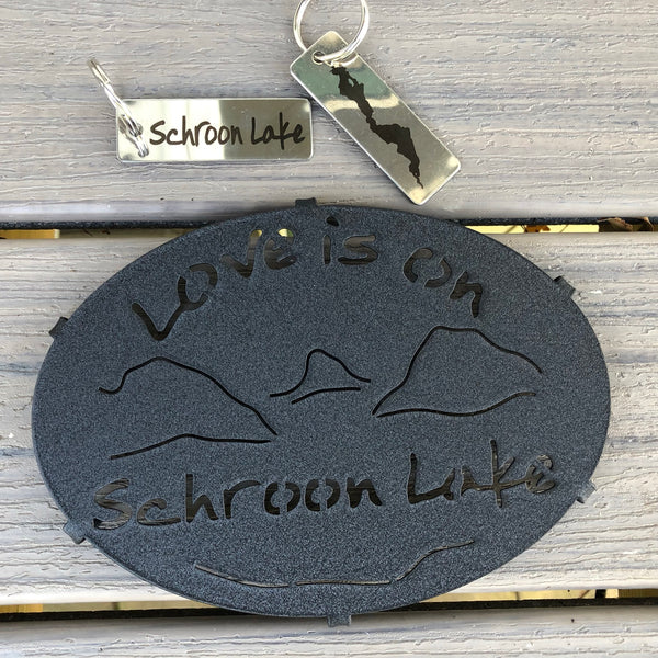 Love is on Schroon Lake text over/under a lake/mountain scene on a black metal trivet. It is paired with a stainless Schroon Lake keychain that has the silhouette of the lake on one side.