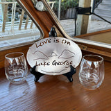 Two Love is on Lake George clear stemless wine glass and a stainless Love is on Lake George trivet.  Both products have our Love is on Lake George lake and mountain scene on them.