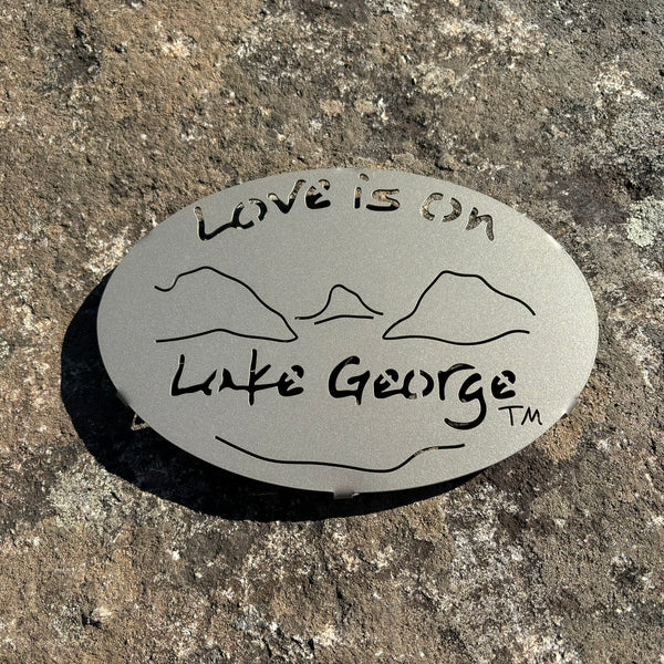 Text Love is on Lake George with the outline of the lake and surrounding Adirondack mountains on a metal trivet; color is gold haze