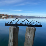 Decorative metal sign with 'Love is in Lake Placid' on a wooden post by a lake.