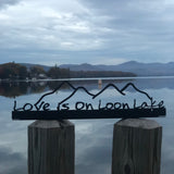 Love is on Loon Lake text with a mountain scape on a black metal sign that sits on a shelf; shelf sitter.