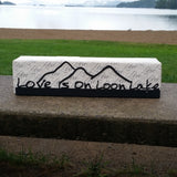 Love is on Loon Lake text with a mountain scape on a black metal sign that sits on a shelf; shelf sitter.