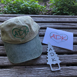 ADK two tone ball cap in khaki/green, ADK Stainless tree shaped bottle opener and and ADK in a heart note card; white background with a green heart and ADK red letters.