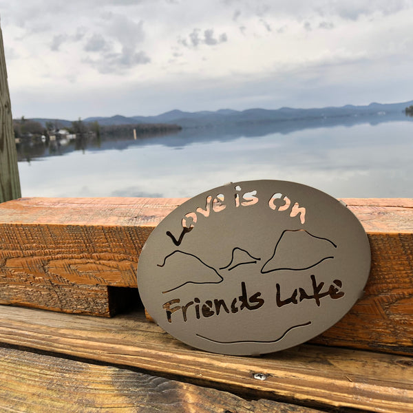 Love is on Friends Lake text with the outline of the lake and surrounding Adirondack mountainscape on a metal trivet; gold haze powder coating