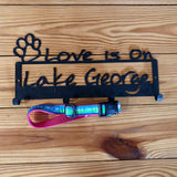Love is on Lake George text on a wall mount with 4 hooks to hang things; paw outline in the upper left corner.  It has a Lake George dog collar hanging from one of the hooks.  It has our Lake George logo in blue and green with a pink backing
