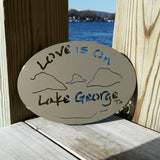 Text Love is on Lake George with the outline of the lake and surrounding Adirondack mountains on a metal trivet; color is gold haze