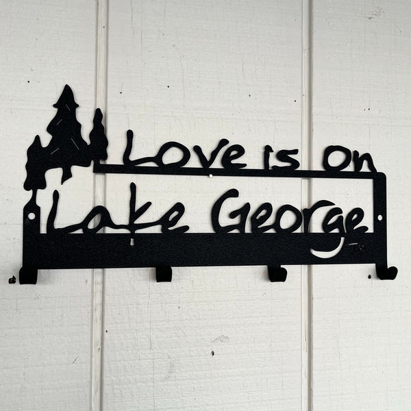 Love is on Lake George text and a cluster of 3 trees cut out on a metal wall mount with 4 hooks for hanging hats, leashes, keys, etc; black