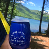 Love is on Lake George text over our lake and mountain scene printed on a 15 oz  stemless plastic wine tumbler or cocktail classic glass.  Text is white; cup is lake blue.