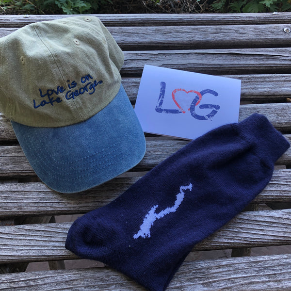 Love is on Lake  George text on a two-tone ball cap, a pair of navy blue socks with the silhouette of Lake George stitched in white, LheartG notecard ; red heart in LG interlocking on a white background.