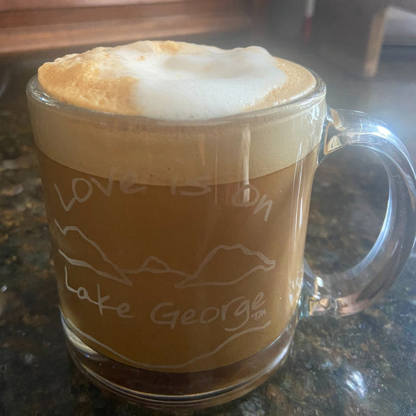 Love is on Lake George text with the outline of the lake and surrounding Adirondack mountains on a clear glass coffee mug.