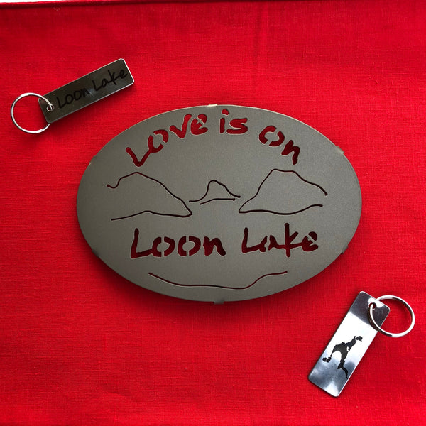 Love is on Schroon Lake text over/under a lake/mountain scene on a black metal trivet.  It is paired with a stainless Schroon Lake keychain  that has the silhouette of the lake on one side. 