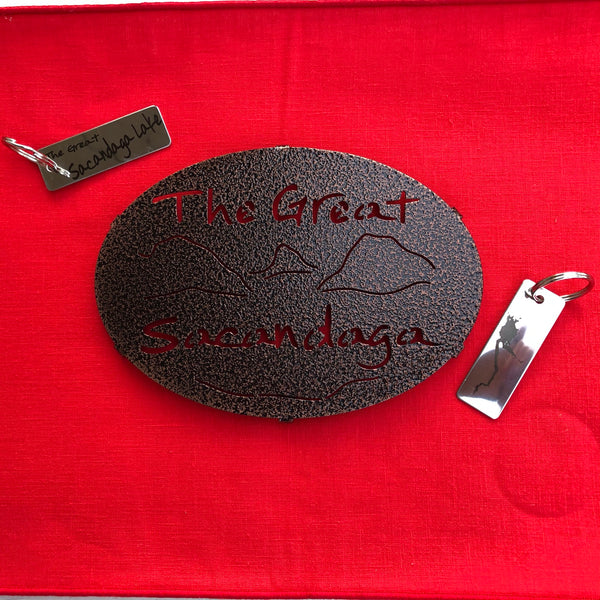 The Great Sacandaga text over/under a lake/mountain scene on a black metal trivet.  It is paired with a stainless Sacandaga keychain that has the silhouette of the lake on oneside. 