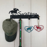 Jockey riding a horse with the word Saratoga.  It is a metal 4 hook wall mount color black/graphite.  A ball cap, dog collar and two horse ornaments are hanging off of the hooks.