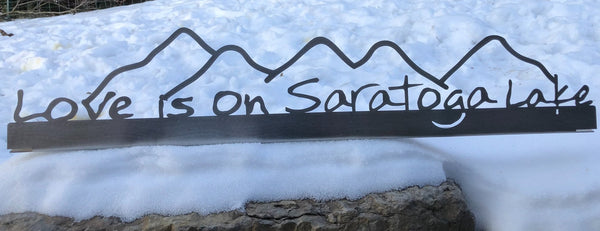 Stainless metal sign with mountains and the text "Love is on Saratoga Lake under the mountains.