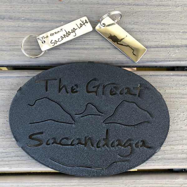 The Great Sacandaga text over/under a lake/mountain scene on a black metal trivet.  It is paired with a stainless Sacandaga keychain that has the silhouette of the lake on oneside. 