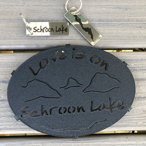 Love is on Schroon Lake text over/under a lake/mountain scene on a black metal trivet.  It is paired with a stainless Schroon Lake keychain  that has the silhouette of the lake on one side. 